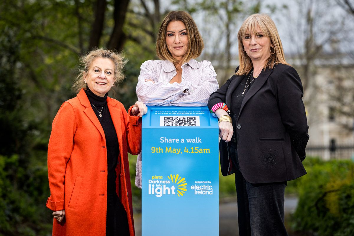 Three women stand beside a "Darkness into Light" event sign, inviting participation on 9th May at 4:15 am.